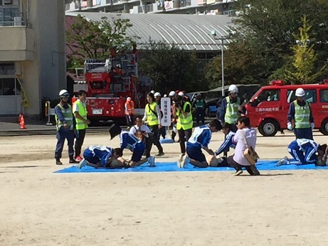 ブルーシートの上に練習用人形がおかれ、2人1組で心肺蘇生法の実演を行っている青いジャージと白いゼッケンを着用した小中学生と、それを監督している黄色いゼッケンをつけた消防隊員たちの写真