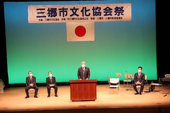 「三郷市文化協会祭」と書かれた横断幕と国旗が掲げられた舞台で、三郷市長が演台に立ち挨拶をし、左右に関係者の方々が座っている写真
