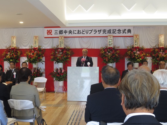 「祝 三郷中央におどりプラザ完成記念式典」と書かれた横断幕と紅白幕、スタンド花が飾られた会場で、関係者と参加者が向かい合って座り、中央の檀上で式辞を述べている市長の写真