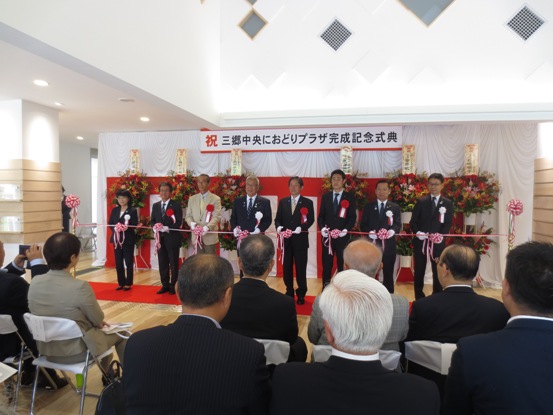 参加者が座っている前で、「祝 三郷中央におどりプラザ完成記念式典」と書かれた横断幕と紅白幕の会場で市長を含め8名の関係者が横一列に並びテープカットを行おうとしている様子の写真