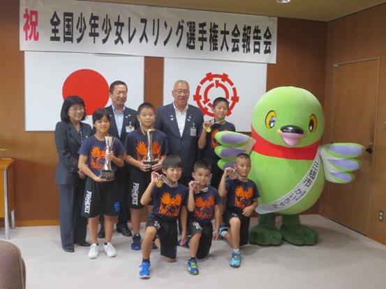 「祝 全国少年少女レスリング選手権大会報告会」と書かれた横断幕と国旗と三郷市旗が飾られた室内でマスコットキャラクターかいちゃんが右側に立ち、市長を中央に3名の子どもが首にメダルをかけて手に持って見せ、2名の子どもがトロフィーを持って立ち、男性と女性が並んでいる記念写真
