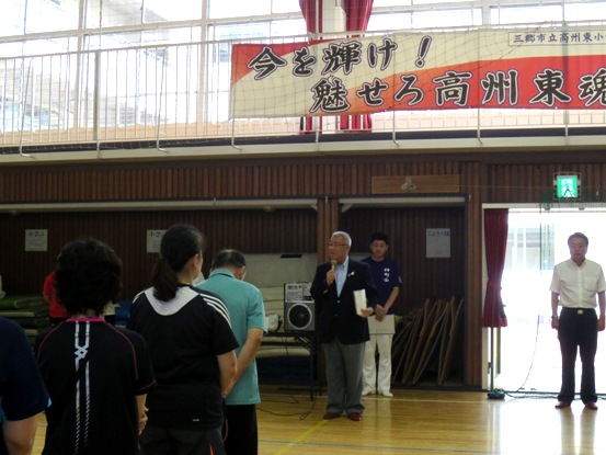 体育館の壁側に2名の関係者が立ち、向かい合って立っている参加者が中央に立ちマイクを持って挨拶を行っている市長の話を聞いている様子の写真