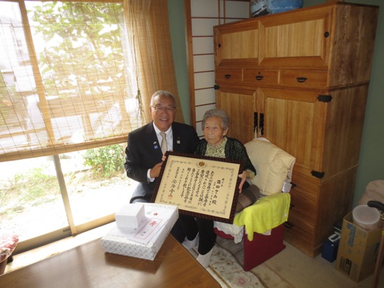 自宅タンス前の椅子に座った篠田さんが、額に入った賞状を持ちその隣に市長が座って記念撮影をしている写真
