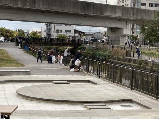 川沿いに設置されたフェンス越しに長い棒を使って川の浮遊ごみを集めている人と、ごみ袋を手に持っている参加者たちの写真