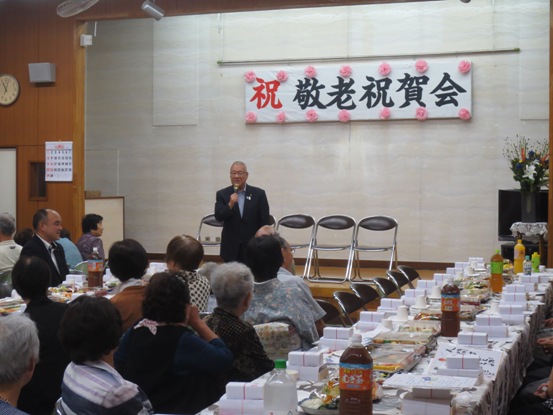 「祝 敬老祝賀会」と書かれた横断幕が貼られたステージ前の席に参加者が座り、右手にマイクを持って挨拶をする市長を見ている大広戸町会の敬老会の様子の写真