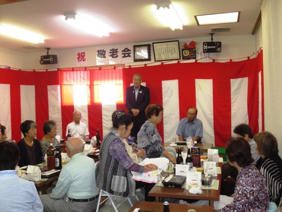 「祝 敬老会」と書かれた文字と紅白幕が貼られた戸ヶ崎一丁目下町会の敬老会の会場で、参加者の前で挨拶を行っている市長の写真