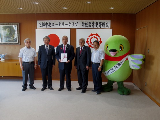 三郷中央ロータリークラブ 学校図書費寄贈式」と書かれた横幕と国旗と三郷市旗が飾られた室内の右側にマスコットキャラクターかいちゃんが立ち、中央に目録を持った市長と4名の男性が横一列に並んでいる記念写真