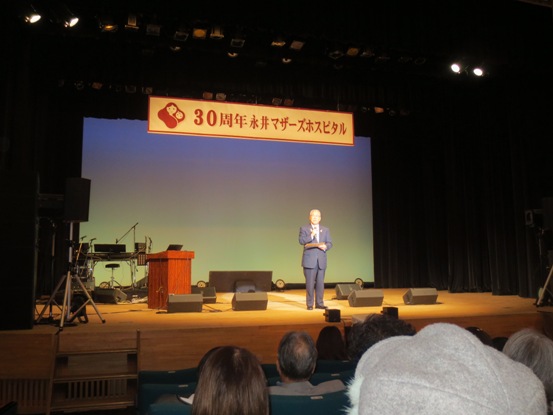 「30周年永井マザーズホスピタル」と書かれた横断幕下のステージ中央に立ち挨拶を行っている市長の写真