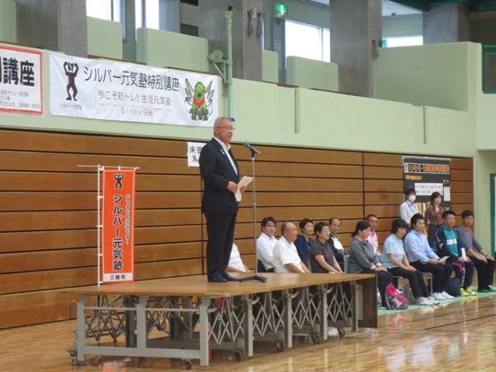 「シルバー元気塾特別講座」と書かれた横幕が壁に貼られた体育館に設置され並べた椅子に関係者が座り、手前の可動式のステージの上で挨拶を行っている市長の写真