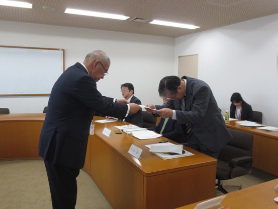 委員の方々が曲線を描いた机の席に座り、手前の席で立ち上がっている男性に市長が委嘱書を交付している様子の写真