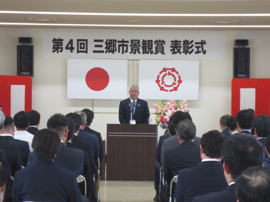 「第4回三郷市景観賞表彰式」と書かれた横断幕と国旗と三郷市旗、紅白幕が貼られた室内の演壇で挨拶を行っている市長の話を、パイプ椅子に座っている参加者が聞いている後ろ姿の写真