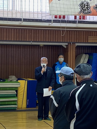 高州東小学校体育館でマイクを持って挨拶している三郷市長と、立って聞いている参加者の方々の写真