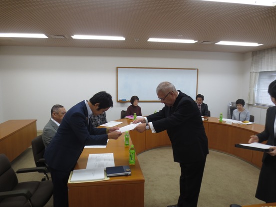 曲線を描いた机の手前の席の男性が立ち、市長から委嘱書を交付されている様子の写真