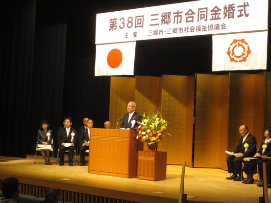 「第38回三郷市合同金婚式」と書かれた横断幕と国旗と三郷市旗が飾られ、金屏風が立てかけられたステージ左右に関係者が座り、中央の演壇で挨拶を行っている市長の写真