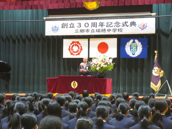 「創立30周年記念式典 三郷市立瑞穂中学校」と書かれた横断幕が飾られたステージで祝辞を述べている市長の話を整列して聞いている生徒たちの後ろから全体を写した写真