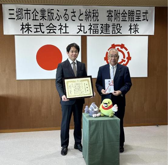 三郷市企業版ふるさと納税 寄附金贈呈式 株式会社丸福建設様の横断幕を背景に左に取締役松本大輔様、右に市長が立っている写真