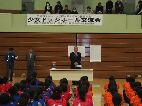 「少女ドッジボール交流会」と書かれた横幕が壁に貼られた体育館の床に体育座りしている子供たちが、前に立っている市長の話を聞いている挨拶の様子の写真