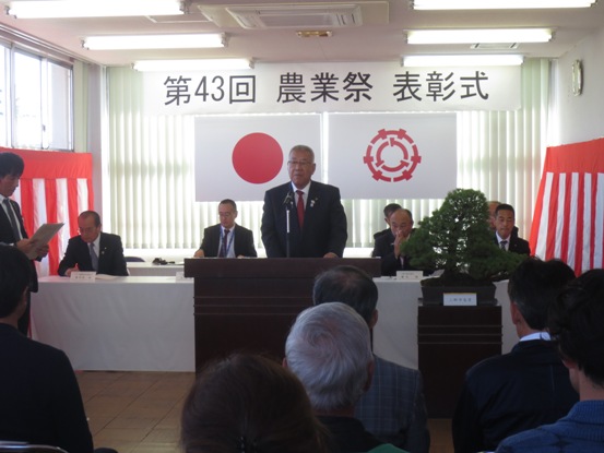 「第43回農業祭表彰式」と書かれた横幕と国旗と三郷市旗、紅白幕が貼られた室内で、関係者と参加者が向かい合って座り、中央の演壇で挨拶を行っている市長を正面から写した写真