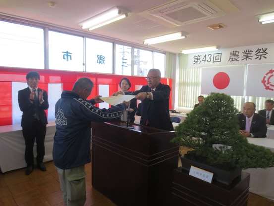 演壇を挟んで立つ男性に市長が賞状を手渡している表彰式の様子の写真