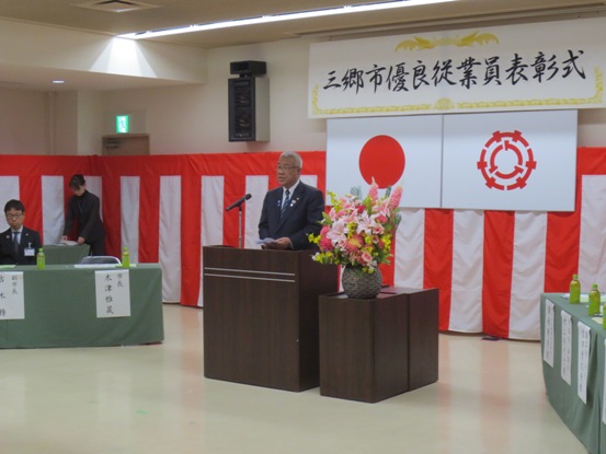「三郷市優良従業員表彰式」と書かれた横断幕と国旗と三郷市旗、紅白幕が貼られた会場の花が飾られた演壇に立ち挨拶を行っている市長の写真