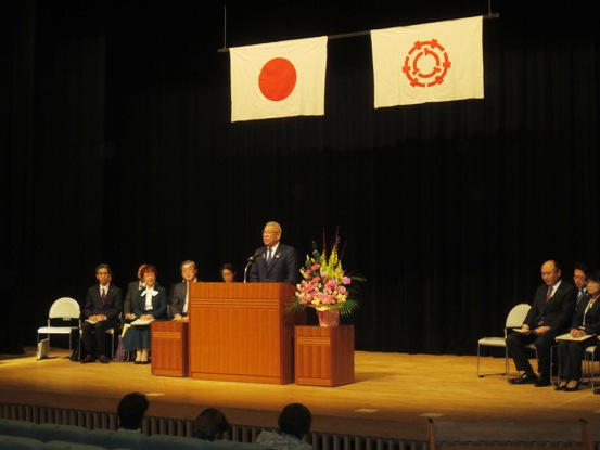 国旗と三郷市旗が飾られたステージ左右に設置された椅子に関係者が座り、中央の演壇で挨拶を行っている市長の写真