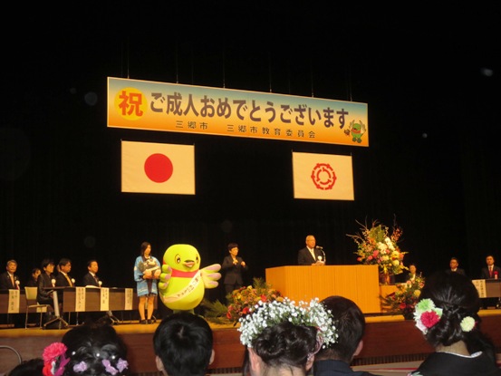 「祝 ご成人おめでとうございます。」と書かれた横断幕と国旗と三郷市旗が飾られたステージ左右の席に関係者が座り、左側にかいちゃんと水色の法被を着た女性がつぶちゃんのぬいぐるみを持って並んで立ち、中央の演壇で挨拶を行っている市長の写真