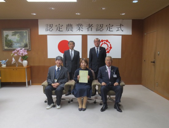 「認定農業者認定式」と書かれた横幕と国旗と三郷市旗が飾られた室内で、前列の椅子に市長と岡永一雄様、認定書を持った浩菜様が座り、後列に2名の男性が立っている写真