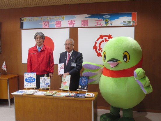 「三郷ウエンズデーロータリークラブ様 図書寄贈式」と書かれた横幕と国旗と三郷市旗が飾られた室内の右側にマスコットキャラクターかいちゃんが立ち、赤色のジャンパーを着た男性と目録を持った市長が横に並び、手前に設置された台の上に表紙が見えるように寄贈された図書が並んでいる写真