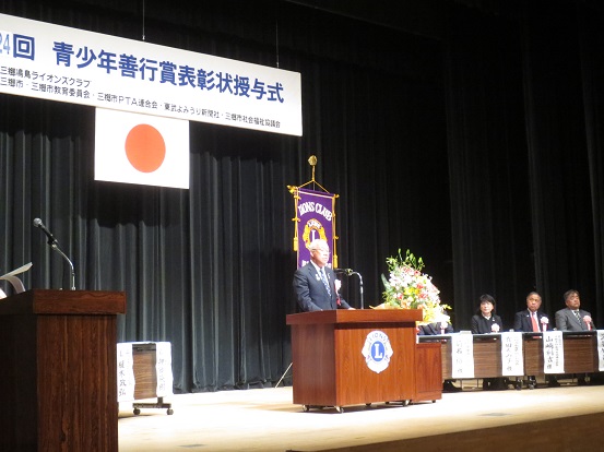 「第24回青少年善行賞表彰状授与式」と書かれた横断幕や国旗が飾られたステージ右側に関係者が座り、中央の演壇で挨拶を行っている市長の写真