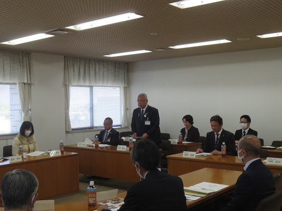 2列に設置された席から立ち上がった市長が挨拶を行い、着席した関係者や委員の方々が説明を聞いている様子の写真
