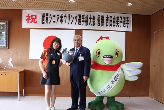 「祝 世界シニアボウリング選手権大会優勝吉田由美子選手」と書かれた横幕と国旗と三郷市旗が飾られた室内右側にマスコットキャラクターかいちゃんが立ち、中央の市長と左に立ちメダルを首にかけている吉田選手が一緒につぶちゃんのぬいぐるみを持っている記念写真