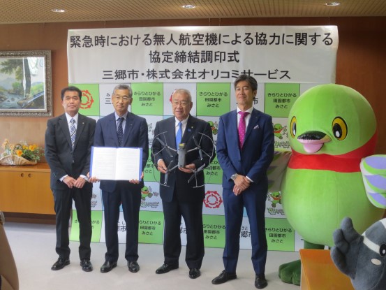 「緊急時における無人航空機による協力に関する協定締結調印式 三郷市・株式会社オリコミサービス」と書かれたバックパネルの前で、マスコットキャラクターかいちゃんが右端に立ち、中央右側にドローンを持っている市長、左側に立っている男性が協定書を持ち、左右に2名の男性が並んで立っている写真