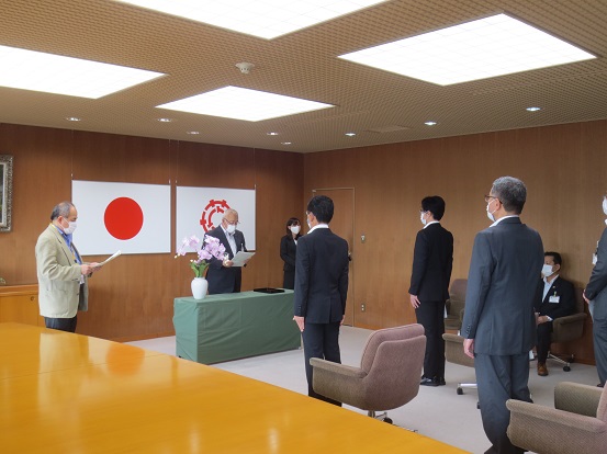 国旗と三郷市旗が飾られた室内で、4名の男性が椅子から立ち上がり、薄緑色のクロスがかけられた台を挟んで向かい合って立っている市長が訓示を述べている様子の写真