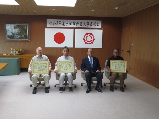「令和2年度三郷市優秀工事表彰式」と書かれた横断幕と国旗・三郷市旗が吊り下げられている壁の前で、三郷市長と表彰状を手に持った3名の受賞者の男性が椅子に座り記念撮影をしている写真