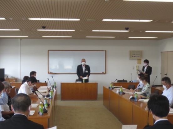 ホワイトボードの前に配置された席に立ち挨拶の言葉を述べる三郷市長と、マイクと席札、ペットボトルのお茶が置かれている席に座り話を聞く委員のメンバーの写真