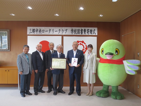 「三郷中央ロータリークラブ学校図書費寄贈式」と書かれた横断幕と国旗・三郷市旗が吊り下げられている壁の前で、お祝儀袋を持った三郷市長とメンバーのうち1人が感謝状を持っている三郷中央ロータリークラブの関係者がかいちゃんとともに記念撮影をしている写真