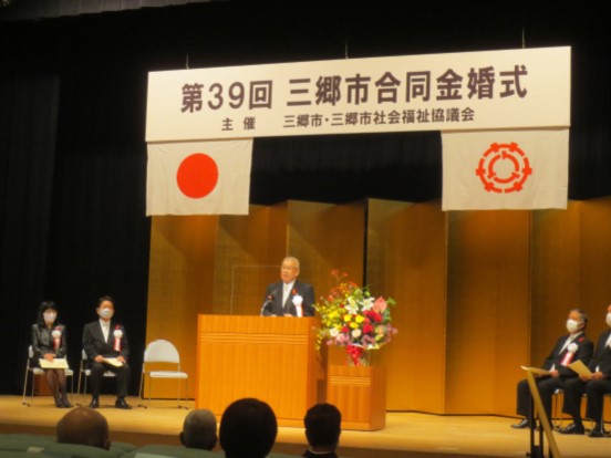 「第39回三郷市合同金婚式」と書かれたボードと国旗・三郷市旗が吊り下げられ、金屏風が置かれているステージ上で、赤・黄色・ピンク・白の豪華な花々で彩られた花台と並ぶ演台に立ち挨拶の言葉を述べる三郷市長と、ステージ上に置かれたパイプ椅子に座り話を聞く胸章を付けた来賓者の写真
