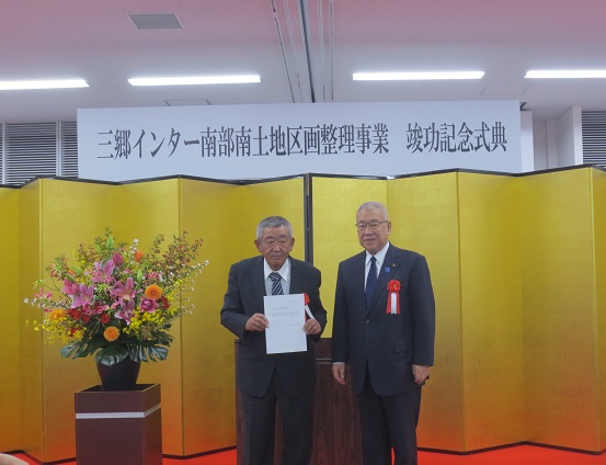 「三郷インター南部南土地区画整理事業竣功記念式典」と書かれたボードが天井から吊り下げられた下に金屛風が設置され、赤・ピンク・オレンジ・黄色・白の豪華な花々で彩られた花台と並ぶ演台の前で、解散認可書を持った男性と三郷市長が記念撮影をしている写真