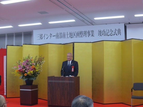 「三郷インター南部南土地区画整理事業竣功記念式典」と書かれたボードが天井から吊り下げられた下に金屛風が設置され、赤・ピンク・オレンジ・黄色・白の豪華な花々で彩られた花台と並ぶ演台に立ち祝辞を述べる三郷市長の写真