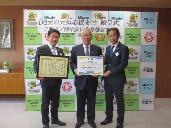 「地元の元気応援寄付贈呈式」と書かれた横幕が貼りつけられている三郷市のバックボードの前で、感謝状を持った常峰越谷支社長、目録をともに持つ木津市長と楠田三郷営業部長が記念撮影をしている写真