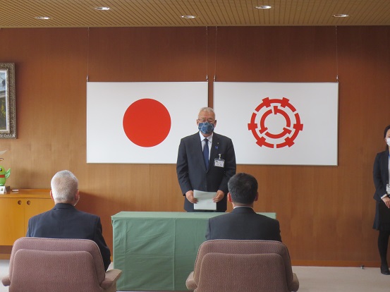 国旗と三郷市旗が吊り下げられている壁の前で、緑のカバーが掛けられた台の後ろに立ち資料を手に持って挨拶の言葉を述べる三郷市長と、椅子に座り話を聞く委員の男性2名の写真