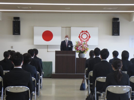 国旗と三郷市章が掛けられた会場で、ピンクや黄色の花が飾られた演台に立って挨拶している三郷市長と、パイプ椅子に座って聞いている参加者の方々の写真