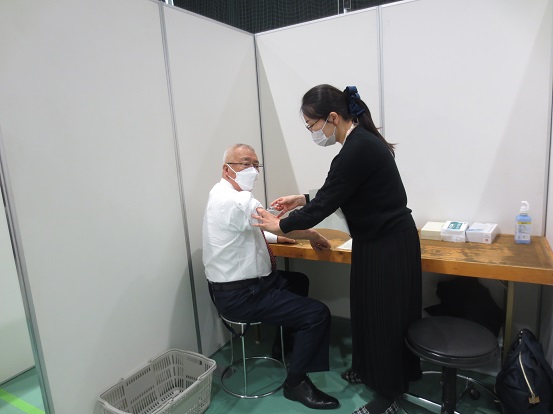 椅子に座ってシャツの右腕をまくり腕を出す三郷市長と、女性スタッフの方がワクチンを打とうとしている写真