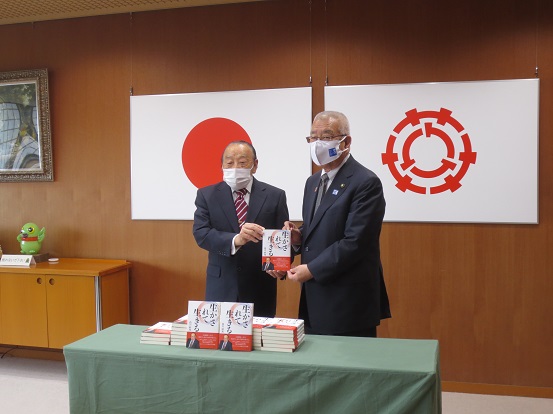 国旗と三郷市章が壁に掛けられた部屋で、くすんだ緑色のカバーが敷かれた机に寄贈された30冊の本を置き、白川好光様と三郷市長が一緒に記念撮影をしている写真