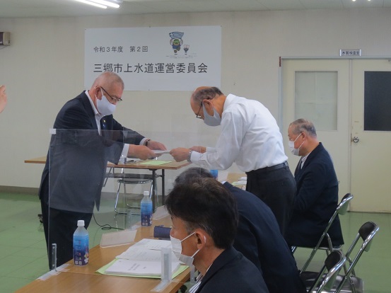 「令和3年度第2回三郷市上水道運営員会」と書かれた横断幕が掲げられた部屋で、三郷市長が委嘱書を男性に手渡ししている写真