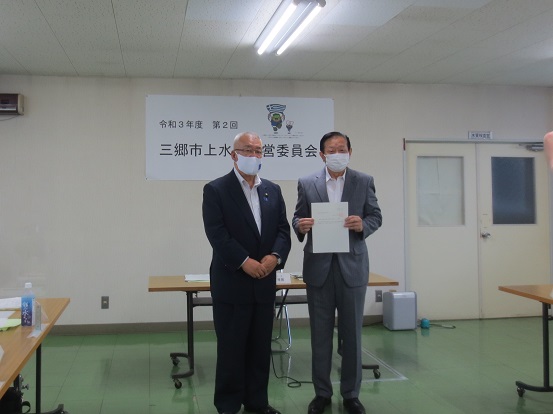 「令和3年度第2回三郷市上水道運営員会」と書かれた横断幕が掲げられた部屋で三郷市長と男性が立ち、男性が書類を手に持ち一緒に記念撮影をしている写真