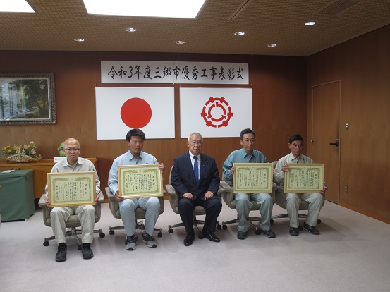 「令和3年度三郷市優秀工事表彰式」と書かれた横断幕と国旗、三郷市章が壁に掛けられた部屋で、賞状を持った4人の男性と三郷市長が椅子に座り記念撮影をしている写真