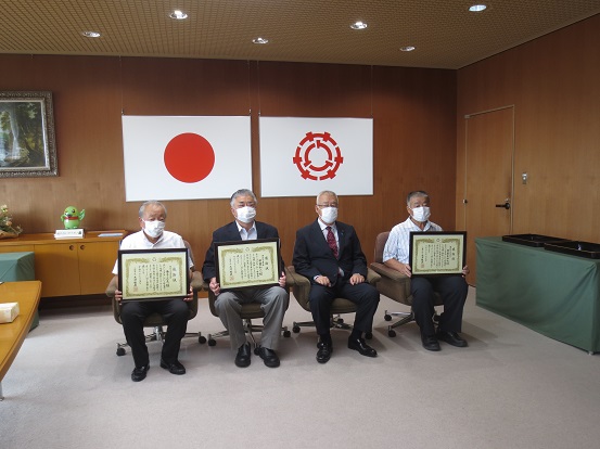 国旗、三郷市章が壁に掛けられた部屋で、感謝状を持った3人の男性と三郷市長が椅子に座り記念撮影をしている写真