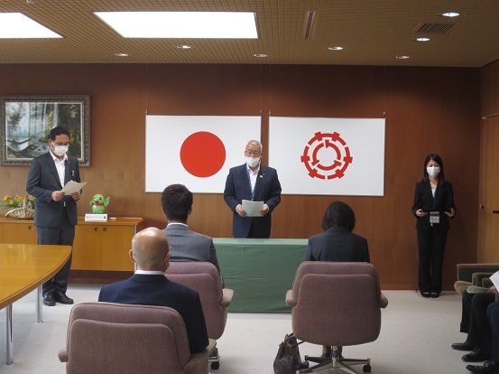 国旗、三郷市章が壁に掛けられた部屋で三郷市長が挨拶をし、左側に男性、右側に女性が立ち、他の関係者の方々は椅子に座っている写真