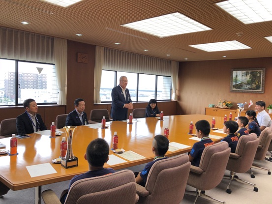 大きな横長のテーブルに男性と6名の子どもたちが座り、向かい合った席から立ち上がり挨拶を行っている市長の写真
